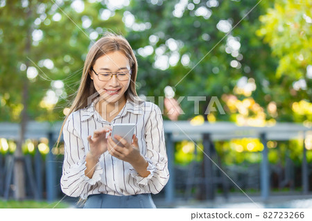 Asian business woman happy smiling looking at smartphone standing park outdoor green nature background 82723266