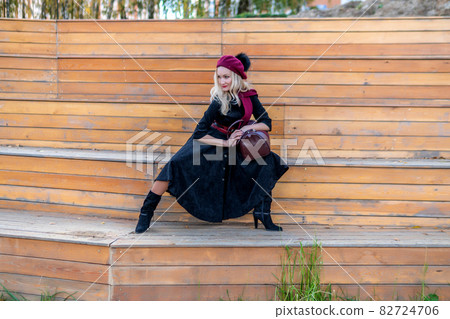 A beautiful woman sits on a summer theater bench, made of wood, in a burgundy coat and biret, with tender lips, in the fall against a background of blue clouds. 82724706