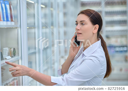 Woman pharmacist consulting by smartphone near shelves with medicines Woman pharmacist consulting by smartphone near shelves with medicines 82725916