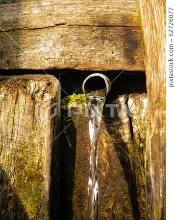 Natural village spring with clean drinking water. Water flows from an iron pipe from a wooden wall. Spring water glare in the sun 82726077