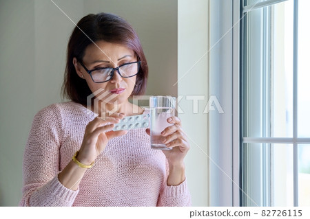 Middle-aged woman with pills and glass of water in her hands, at home Middle-aged woman with pills and glass of water in her hands, at home 82726115