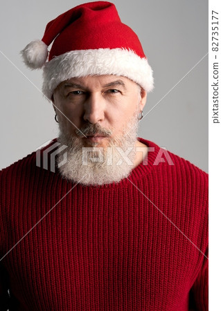 Portrait of trendy middle aged man with beard wearing Christmas hat and red sweater looking at camera while posing isolated over gray background Portrait of trendy middle aged man with beard wearing Christmas hat and red sweater looking at camera while posing isolated over gray background 82735177