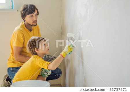 Man with his son makes repairs at home, he teaches children to plaster the walls with a spatula in his hands 82737371