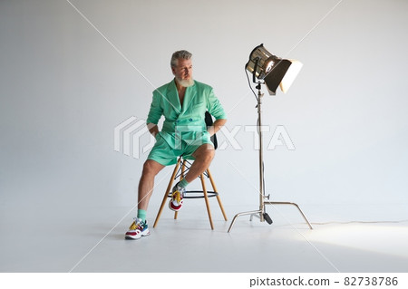 Studio shot of cool gray haired mature man wearing stylish outfit with colorful sneakers looking aside while sitting next to studio spotlight, posing for camera over white background 82738786