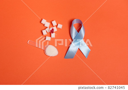 Pure refined white sugar cubes with blood drop and blue satin ribbon, symbol of World diabetes awareness day 14 November. Isolated over colored orange background, copy space for medical ad. Flat lay 82741045