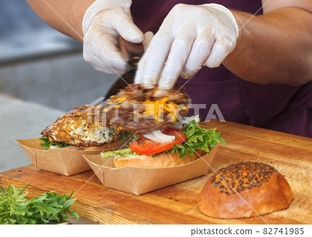 A chef prepares a burger for a customer. A man wearing white protective gloves picks up the burgers from the grill and adds them to the burger bun. Motion blurred burger blade. A chef prepares a burger for a customer. A man wearing white protective gloves picks up the burgers from the grill and adds them to the burger bun. Motion blurred burger blade. 82741985