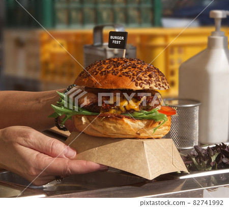 A chef prepares a hamburger for a customer. The woman takes the hamburger box and hands it to the customer. Kitchen working environment in the background. A chef prepares a hamburger for a customer. The woman takes the hamburger box and hands it to the customer. Kitchen working environment in the background. 82741992