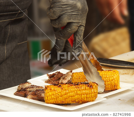 Chef prepares paper trays with grilled food, meat and corn for customers 82741998