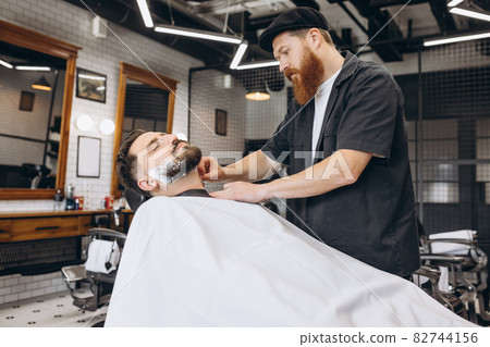 Portrait of professional barber, clipper cutting, shaving beard of regular customer at barbershop. Beauty, selfcare, style, fashion and male cosmetics concept. Portrait of professional barber, clipper cutting, shaving beard of regular customer at barbershop. Beauty, selfcare, style, fashion and male cosmetics concept. 82744156