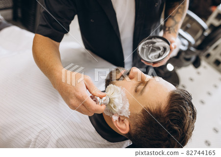 Close-up male hands with shaving brush prepare beard of customer for cutting at barbershop. Beauty, selfcare, style, fashion and male cosmetics concept. Close-up male hands with shaving brush prepare beard of customer for cutting at barbershop. Beauty, selfcare, style, fashion and male cosmetics concept. 82744165