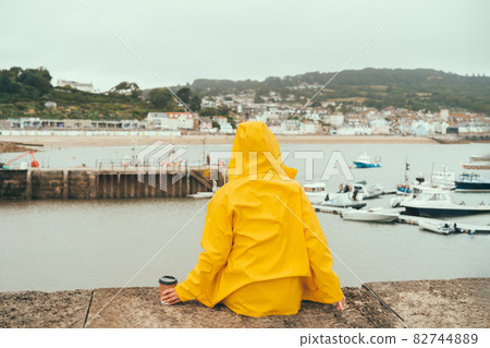 Back view woman in yellow raincoat drinking coffee and enjoying the view of the bay with boats in cloudy weather. Local travel to Lyme Regis, England 82744889