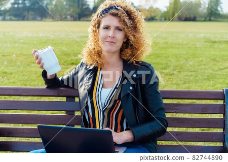 A portrait of woman in casual clothes drinking coffee from reusable cup and using a laptop outdoors sitting on the bench in the public park. Remote work, freelance workplace. Networking. A portrait of woman in casual clothes drinking coffee from reusable cup and using a laptop outdoors sitting on the bench in the public park. Remote work, freelance workplace. Networking. 82744890