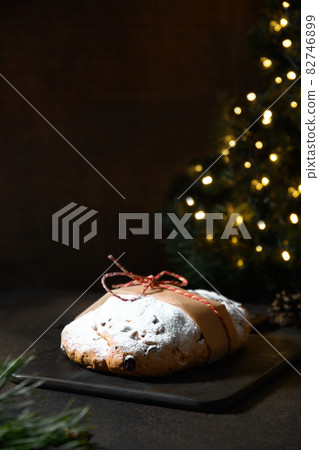 Traditional stollen decorated holiday rope on dark festive table with Christmas tree. Traditional stollen decorated holiday rope on dark festive table with Christmas tree. 82746899