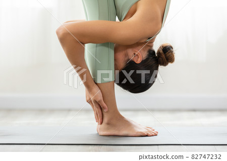 Close up Asian woman wearing green sportwear doing Yoga exercise in front of windows.Yoga Standing Forward Bend pose or Uttanasana.Calm of healthy young woman breathing and meditation yoga at home 82747232