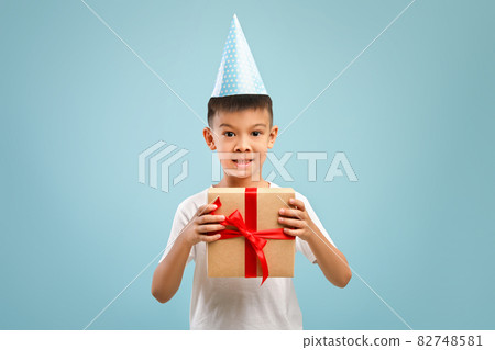 Excited little asian birthday boy wearing party hat holding wrapped gift box 82748581