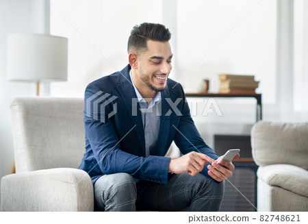 Smiling Arab businessman in formal wear using mobile phone, checking business messages, sitting in armchair at office Smiling Arab businessman in formal wear using mobile phone, checking business messages, sitting in armchair at office 82748621