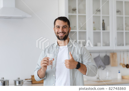 Smiling adult european man with beard holding glass of water and showing thumb up at kitchen interior Smiling adult european man with beard holding glass of water and showing thumb up at kitchen interior 82748860