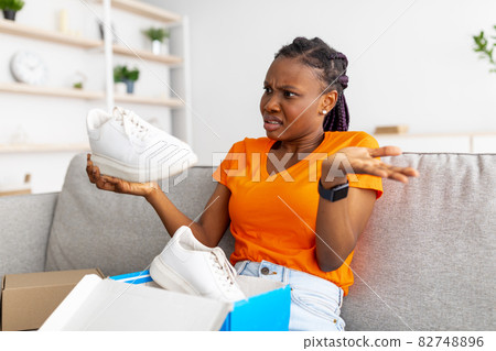 Angry black female opening box from online store and looking at delivered item, upset about received sneaker shoes 82748896