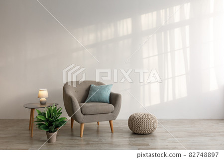 Comfortable armchair with pillow, luminous lamp on table, potted plant and ottoman on floor on gray wall background 82748897