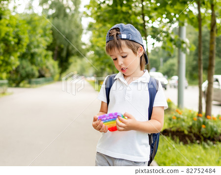 Little boy plays square rubber fidget toy Pop It. Rainbow-colored anti stress and tactile toy for all ages. Outdoor recreation with toy for fine motor skills improvement. 82752484