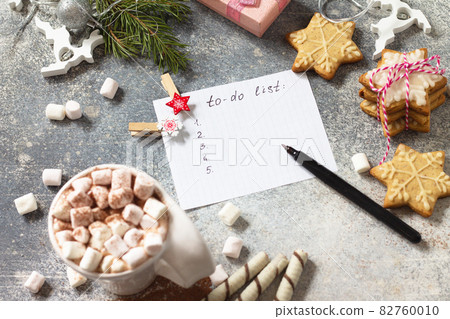 A cup of hot cocoa with marshmallows, Christmas sweets and a to-do list on a stone countertop. A cup of hot cocoa with marshmallows, Christmas sweets and a to-do list on a stone countertop. 82760010