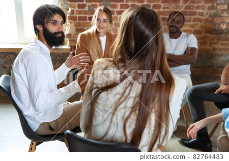Young multiethnic people, men and woman sitting together, talking, communicate at psychological support group, indoors. Young multiethnic people, men and woman sitting together, talking, communicate at psychological support group, indoors. 82764483