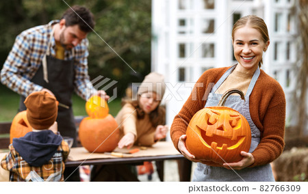Smiling woman with jack-o-lantern smiling at camera while carving pumpkins with family in backyard 82766309