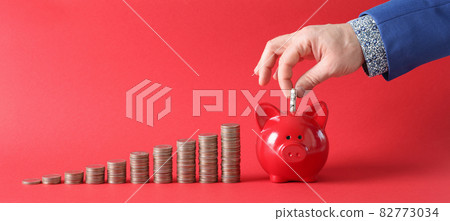 Businessman fills piggy bank with dollar banknote next to there stacks of coins on red background 82773034