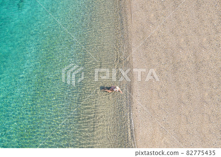 Young woman in hat, swimsuit sitting in the water on the shore of sea on the sand beach. Summer. View from above. Top view, copter 82775435