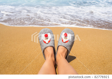 Christmas sea holiday. Woman in santa slippers relaxing on paradise beach island with waves. New year Christmas sea holiday. Woman in santa slippers relaxing on paradise beach island with waves. New year 82776921