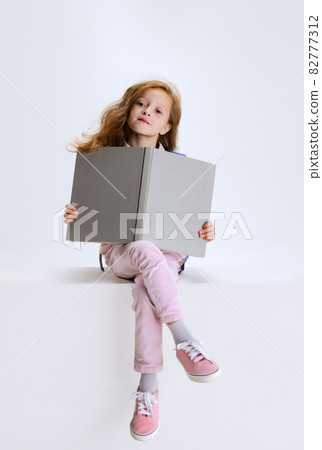 One little cute girl in casual clothes sitting on big box and reading book isolated on white studio background. 82777312