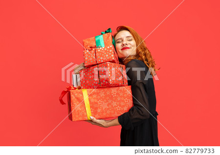 Cropped portrait of beautiful, smiling, redheaded woman holding many gift boxes isolated over red background. Holiday shopping 82779733