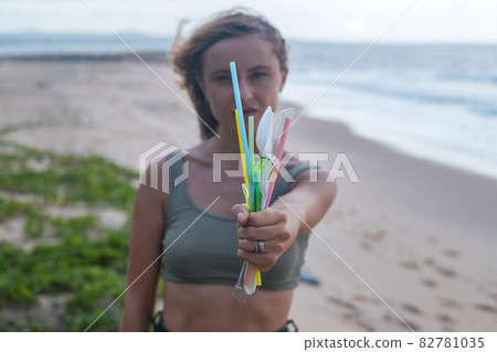 Girl stands on the beach near the ocean and holds used plastic dishes in one hand. Clean ocean beach on the background. Single-use plastic waste  82781035