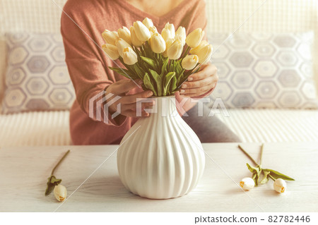 Woman putting white tulips flowers in vase sitting at the living room coffee table. Lifestyle 82782446