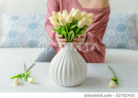 Woman putting white tulips flowers in vase sitting at the living room coffee table. Lifestyle Woman putting white tulips flowers in vase sitting at the living room coffee table. Lifestyle 82782450