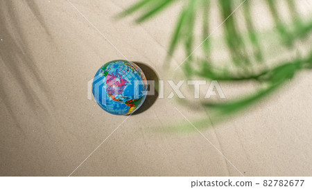 Rubber toy globe on sandy tropical beach under branches of palm and shadow palm tree. Holiday concept. Creative, background, travel, summer. 82782677