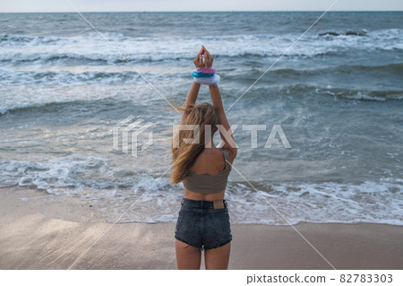 Blond young woman with hands wrapped in blue, pink and white plastic bags on the beach. Back view. Sea on the background. Environment protection. Blond young woman with hands wrapped in blue, pink and white plastic bags on the beach. Back view. Sea on the background. Environment protection. 82783303