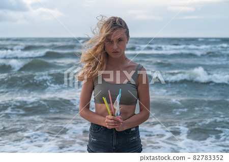 Portrait of Sad blond caucasian young woman standing on the beach and holding used plastic dishes and straws. Sea on the background. plastic pollution 82783352