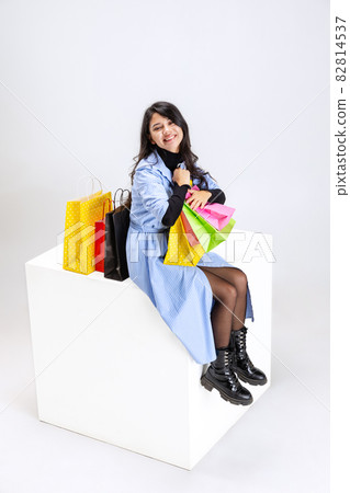 Happy smiling young girl with lots of bags, colorful bright packages sitting on huge box isolated over white studio background. Happy smiling young girl with lots of bags, colorful bright packages sitting on huge box isolated over white studio background. 82814537