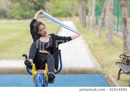Happy smiling young beautiful asian woman in sportswear relaxing on public exercise machine. Cute and sporty girl working out on bike during morning in summer outdoor. 82814890