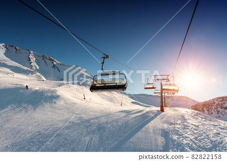 Ski lift empty ropeway on hilghland alpine mountain winter resort on bright sunny evening . Ski chairlift cable way with people enjoy skiing and snowboarding. Sunset sky backlit shining on background 82822158