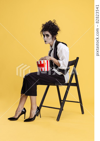 Full-length portrait of creepy looking woman in Halloween costume with popcorn basket sitting on chair isolated on yellow background. Halloween party Full-length portrait of creepy looking woman in Halloween costume with popcorn basket sitting on chair isolated on yellow background. Halloween party 82823541