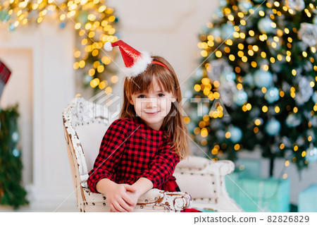 cute little girl in a red plaid dress and a cap in the New Year's interior. cute little girl in a red plaid dress and a cap in the New Year's interior. 82826829