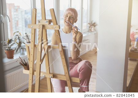 Positive woman with colorful hair bites pencil sitting at easel Positive woman with colorful hair bites pencil sitting at easel 82841045