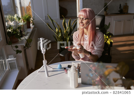 Cheerful woman with glasses applies cream onto palm from bottle shooting tutorial at table Cheerful woman with glasses applies cream onto palm from bottle shooting tutorial at table 82841065