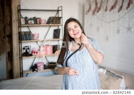 A pregnant girl listens with a phonendoscope to her baby in her belly and smiles. 82842140