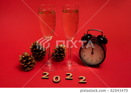 Christmas and New Year concept. Champagne flutes with a sparkling wine, alarm clock showing midnight in the face clock, golden pine cones and wooden numerals 2022, on red background with copy space 82843470