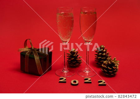 Composition of New year object, wooden 2022 numerals, golden pine cones, Christmas gift in glitter wrapping paper with golden bow, champagne flutes with sparkling wine on red background, copy space 82843472