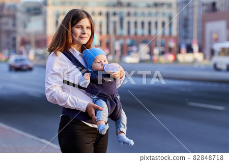 Woman is feeding baby in sling from bottle outdoors. Woman is feeding baby in sling from bottle outdoors. 82848718