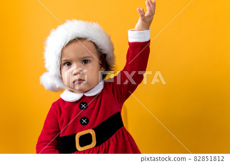 Serious Portraite Cute Happy Cheerful Chubby Baby Girl in Santa Hat Waving Hand Up At Yellow Background. Child Play Christmas Scene Celebrating Birthday. Kid Have Fun Spend New Year Time Copy Space Serious Portraite Cute Happy Cheerful Chubby Baby Girl in Santa Hat Waving Hand Up At Yellow Background. Child Play Christmas Scene Celebrating Birthday. Kid Have Fun Spend New Year Time Copy Space 82851812